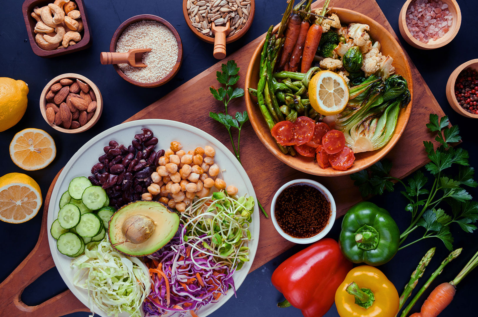 A vibrant display of fresh, colorful vegetables and nuts: two bowls with avocado, cucumbers, chickpeas, and greens; tomatoes, lemon, and spices scattered around.