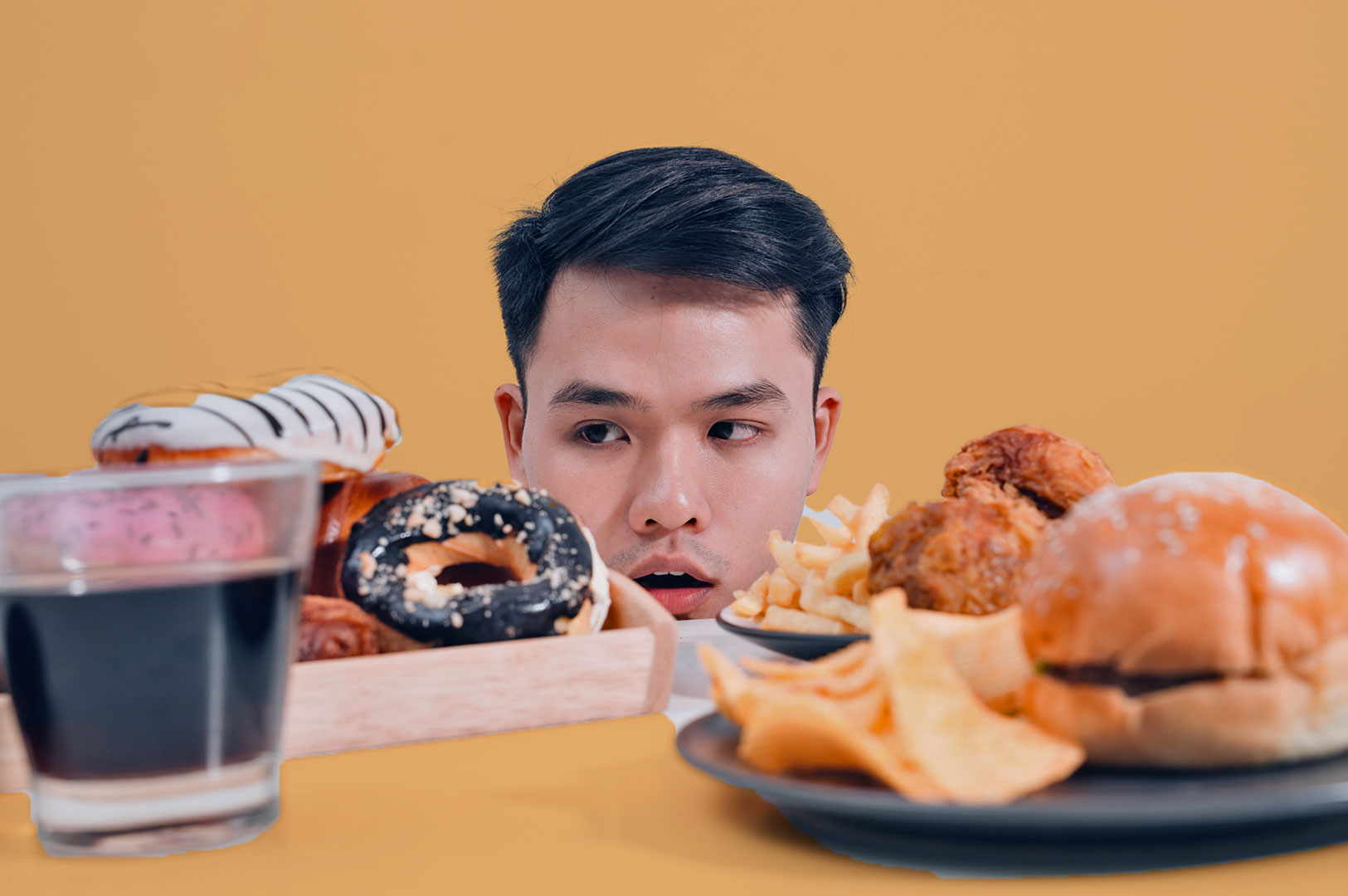 A man with a surprised expression peers over a table filled with donuts, a burger, fries, and a drink against a yellow background. The mood is playful.