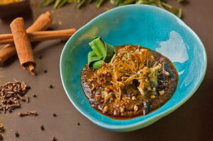 A vibrant blue bowl holds a rich curry garnished with shredded vegetables and herbs. Cinnamon sticks and cloves lie nearby on a brown surface.