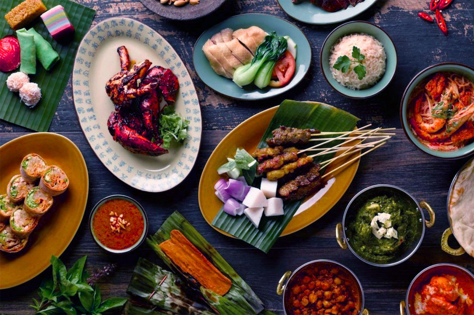An overhead view of assorted Singaporean dishes, including grilled chicken, satay, rice, curries, vegetables, and various sauces, arranged on a rustic wooden table.