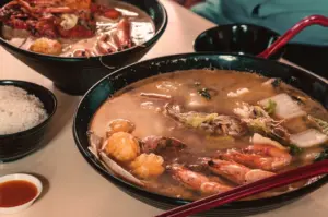 Two bowls of steaming seafood noodle soup with prawns and vegetables on a table. Side of white rice and dipping sauce, creating a warm, inviting meal scene.