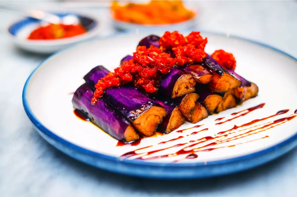 Sliced purple eggplant drizzled with sauce and topped with vibrant red chili paste on a white plate, conveying a spicy and vibrant culinary presentation.