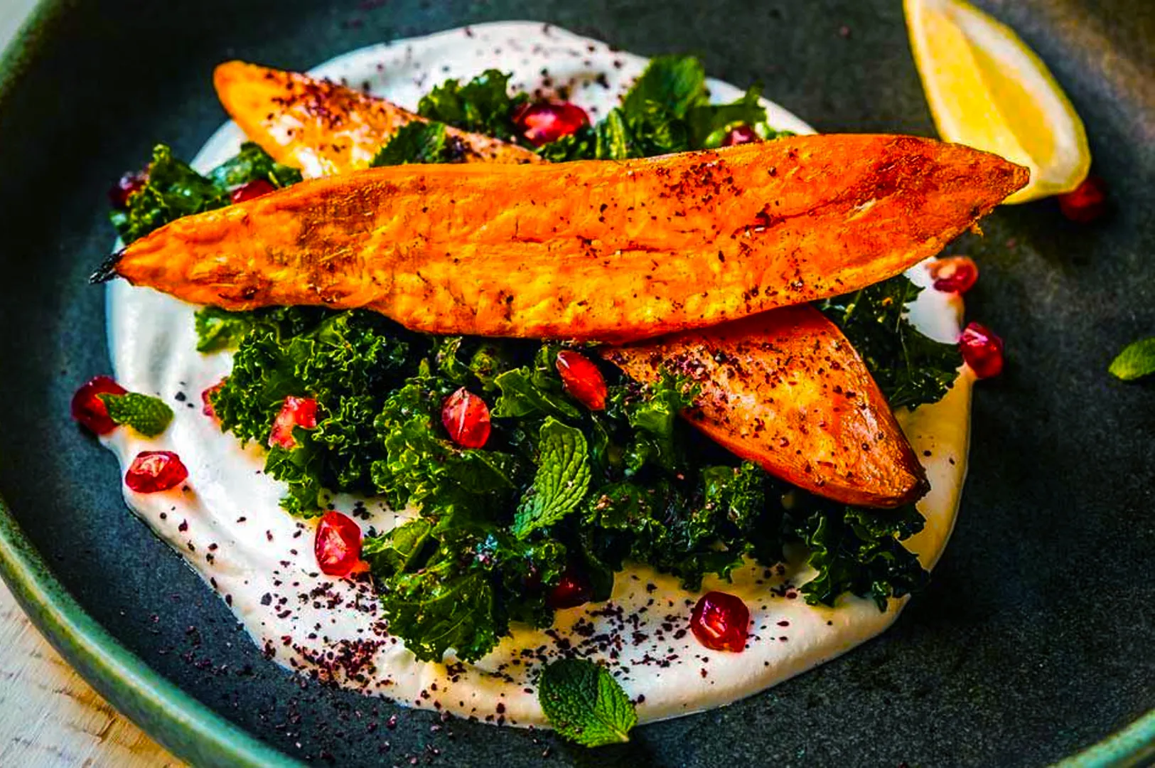 Roasted sweet potato on a bed of kale with creamy white sauce, garnished with pomegranate seeds and mint, lemon wedge on the side. Vibrant and appetizing.