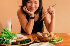 Smiling woman holds chopsticks as she admires a table of Vietnamese dishes, including fresh greens and fried rolls, set against a warm, inviting background.