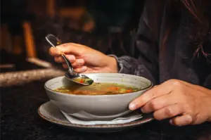 A person holds a spoon above a bowl of vegetable soup, ready to eat. The warm lighting and cozy setting suggest a comforting meal