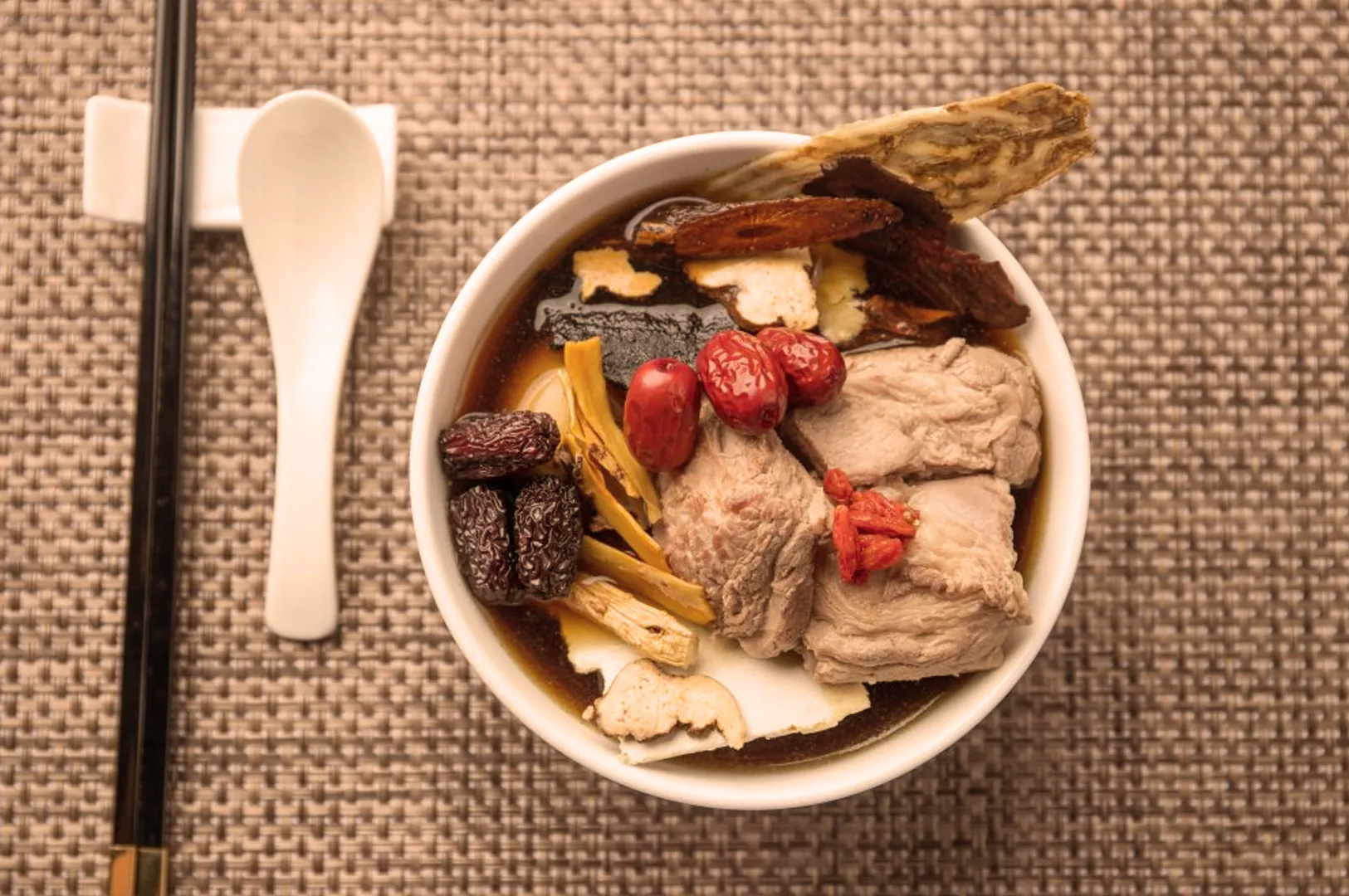 Bowl of herbal soup with meat, red dates, and roots on woven mat. Chopsticks and spoon beside it. Earthy, nourishing ambiance.