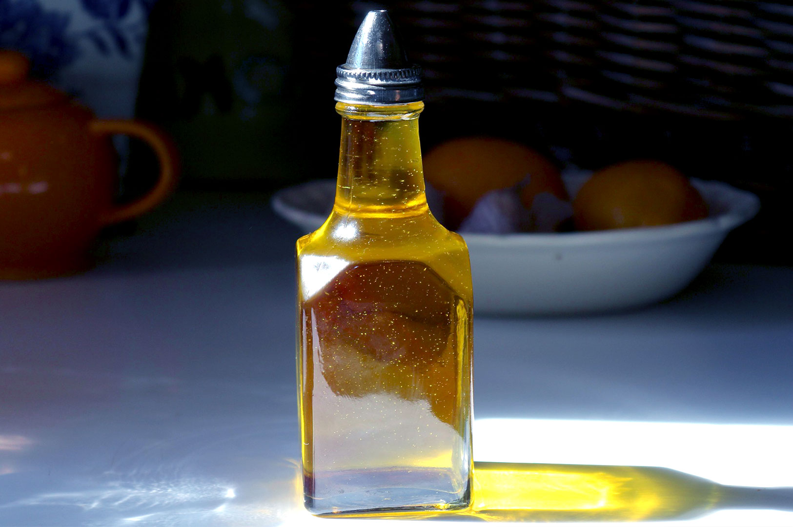 A small glass bottle filled with golden oil sits on a sunlit surface, casting a shadow. Behind it, a blurred bowl of oranges adds a warm, cozy atmosphere.