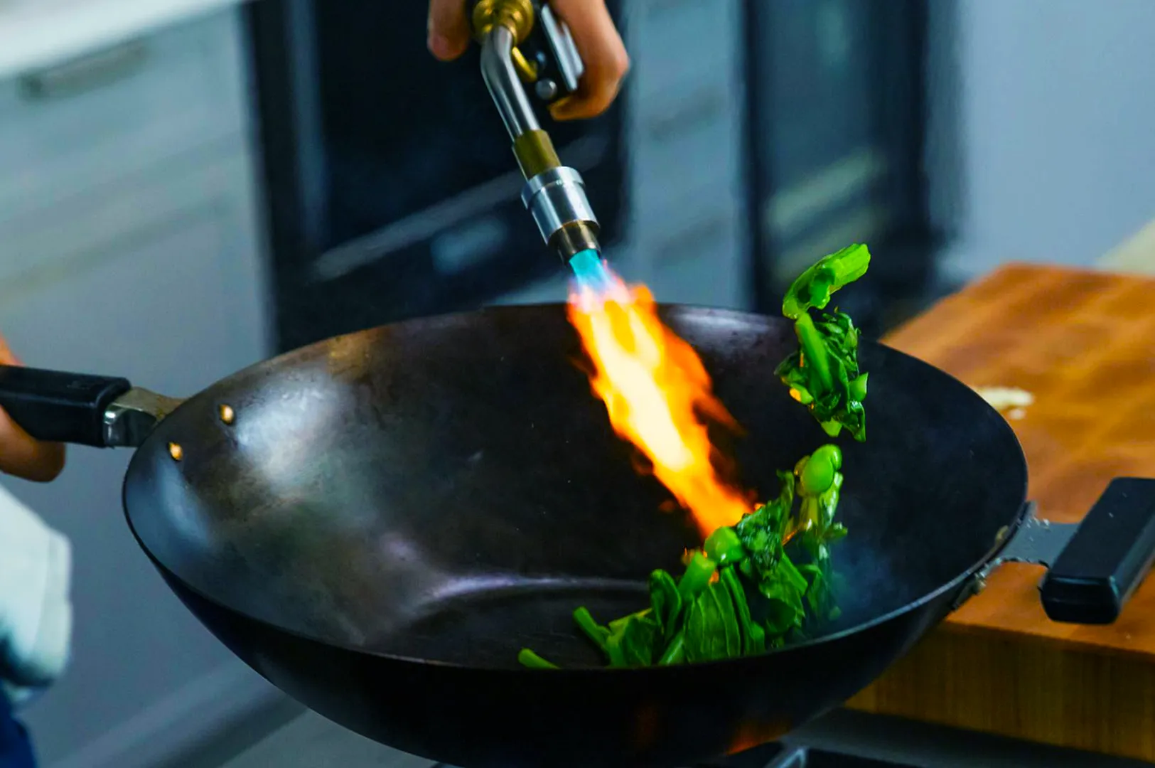 A person uses a blowtorch to cook leafy greens in a black wok, creating a vibrant flame. The scene conveys a dynamic and modern kitchen technique.