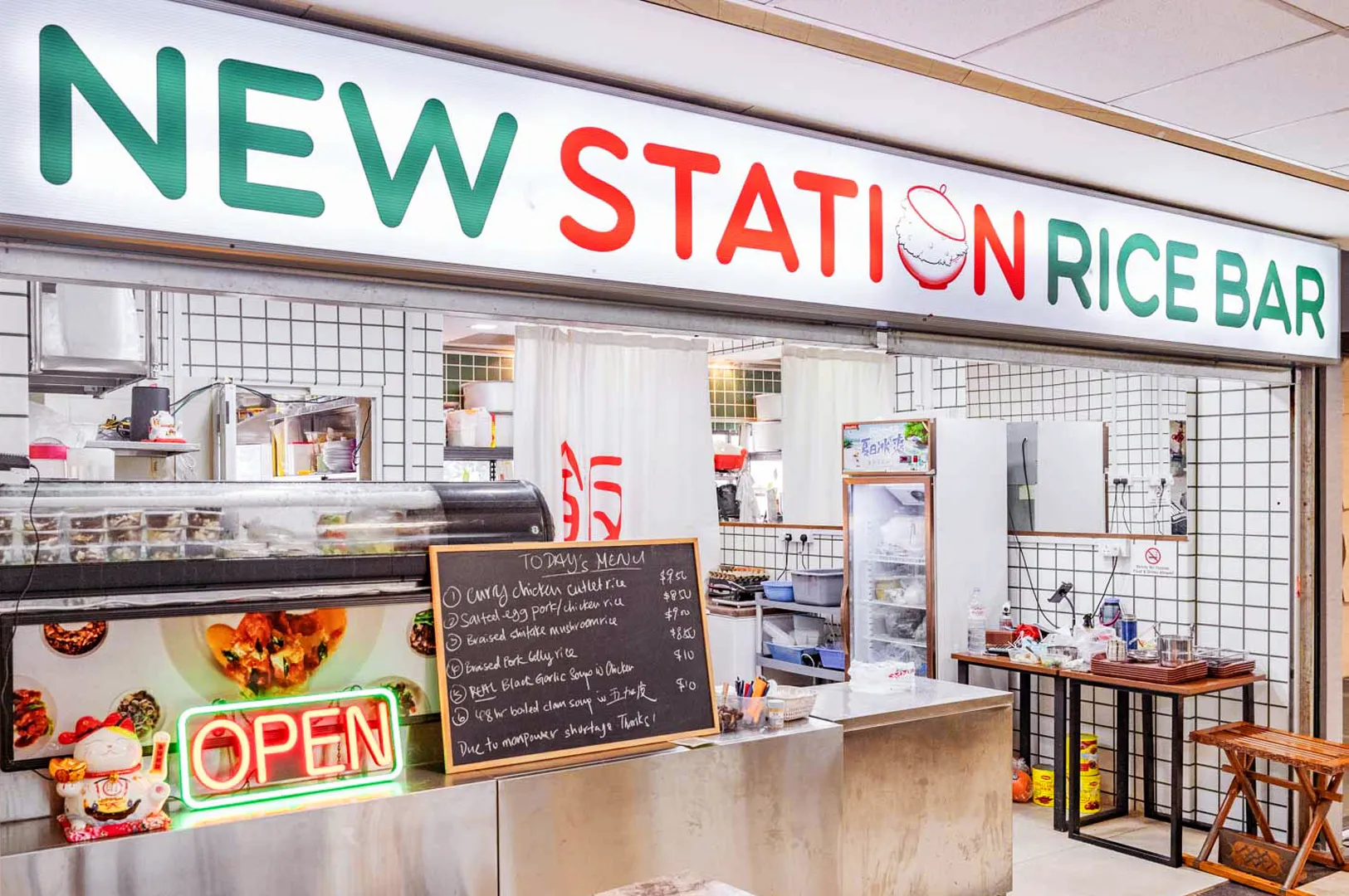 Brightly lit food stall named "New Station Rice Bar" with a neon "Open" sign. A chalkboard displays the menu, creating a welcoming atmosphere.
