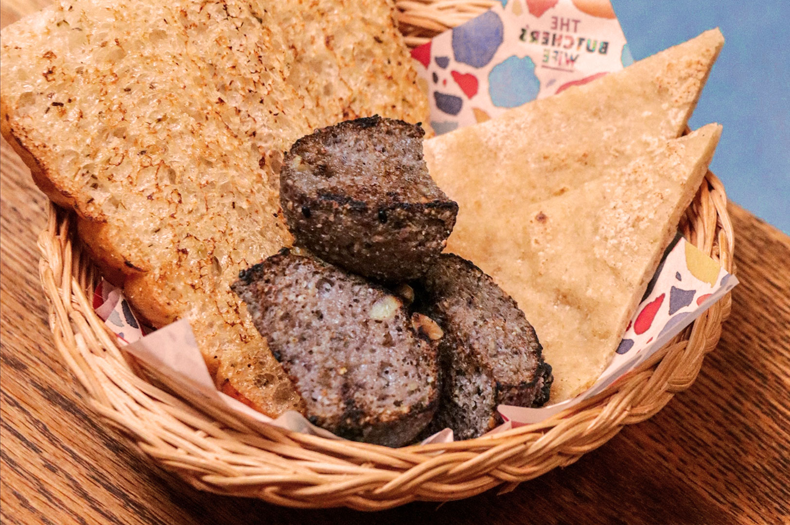 A wicker basket contains sliced black pudding, toasted brown bread, and buttered flatbread on colorful paper, set on a wooden surface.