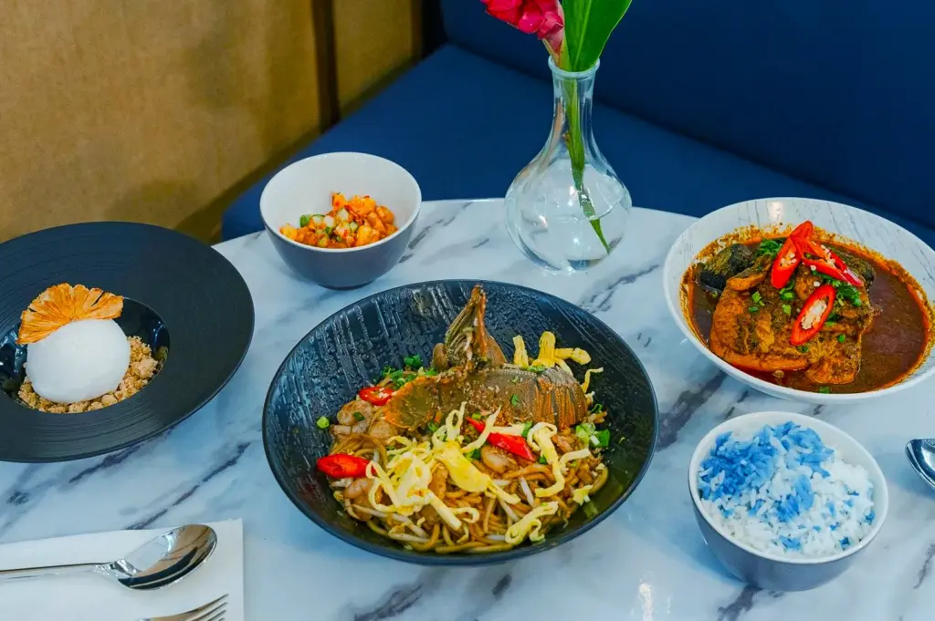 A marble table displays a vibrant meal with spicy noodles, curry with red chili, blue-tinted rice, a white dessert, and a salad, set near a vase with flowers.