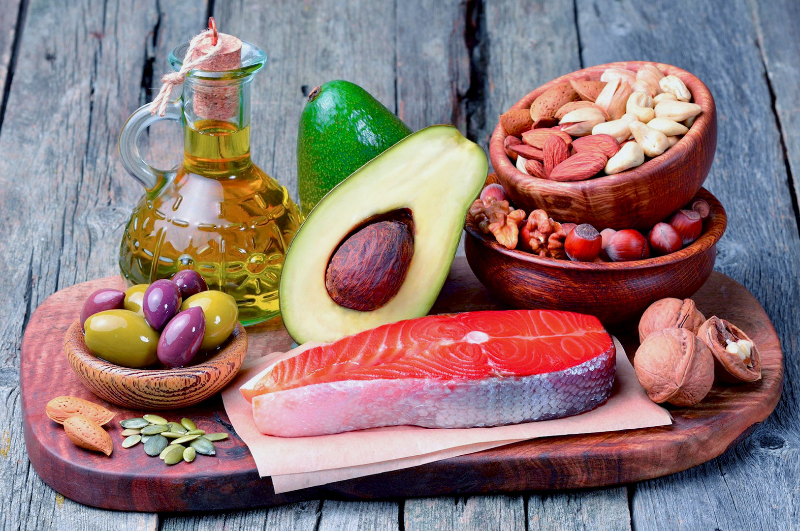 A wooden platter with salmon, avocado, olive oil, nuts, seeds, and olives on a rustic table. Natural, healthy food conveying freshness and nutrition.
