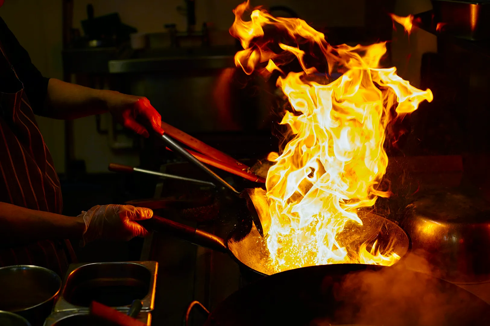 Chef skillfully tossing food in a flaming pan, creating dramatic flames in a dimly lit kitchen. The scene conveys excitement and culinary artistry.