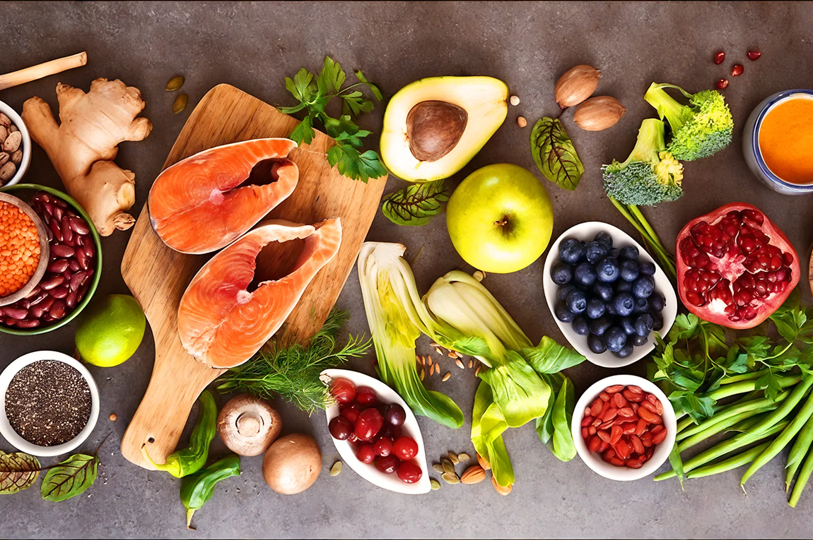 A vibrant spread of healthy foods on a gray surface, including salmon, avocado, blueberries, broccoli, pomegranate, nuts, lentils, and fresh greens.