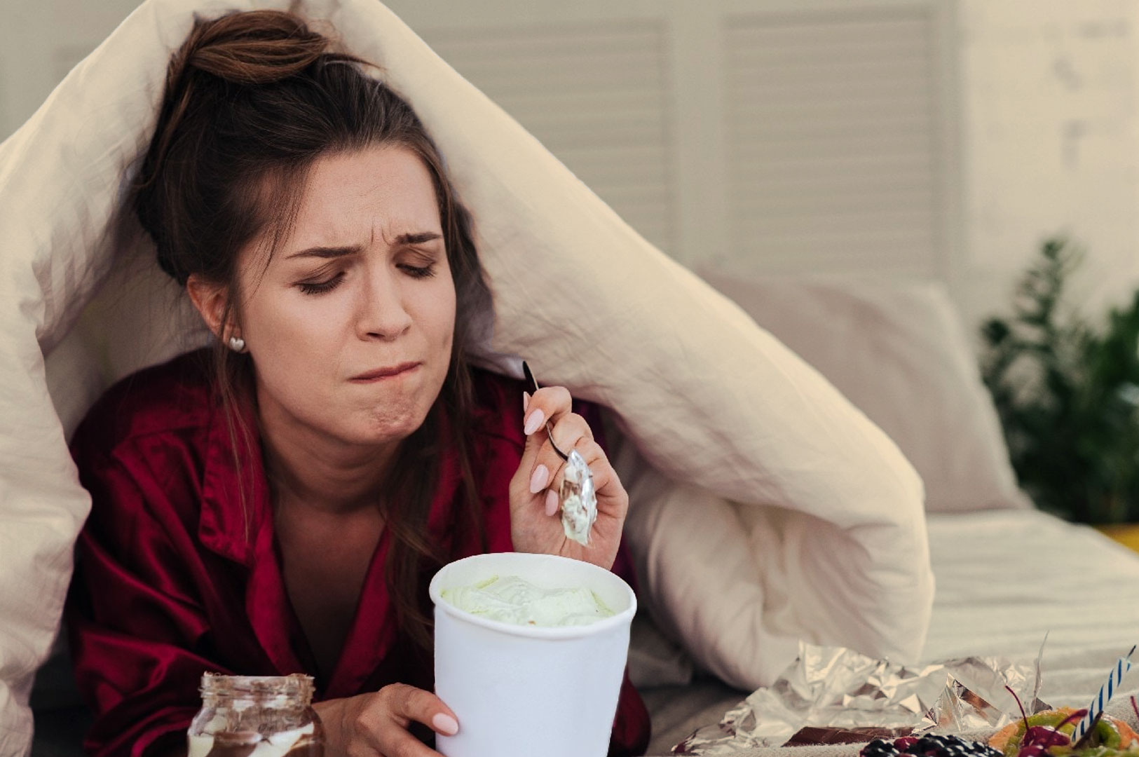 Woman under a blanket in bed, wearing a red robe, holding a spoon and a tub of ice cream. Her expression is emotional, suggesting sadness or frustration.