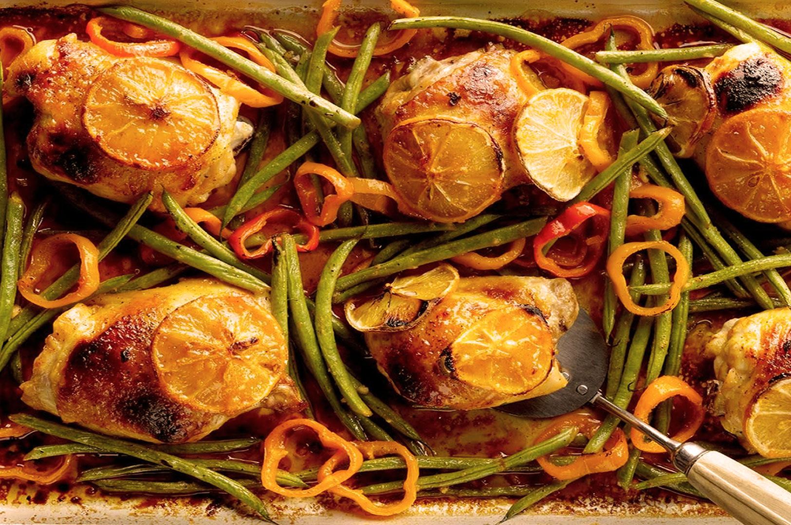 Roasted chicken thighs topped with lemon slices, surrounded by green beans and orange bell pepper rings on a baking sheet. Vibrant and savory.