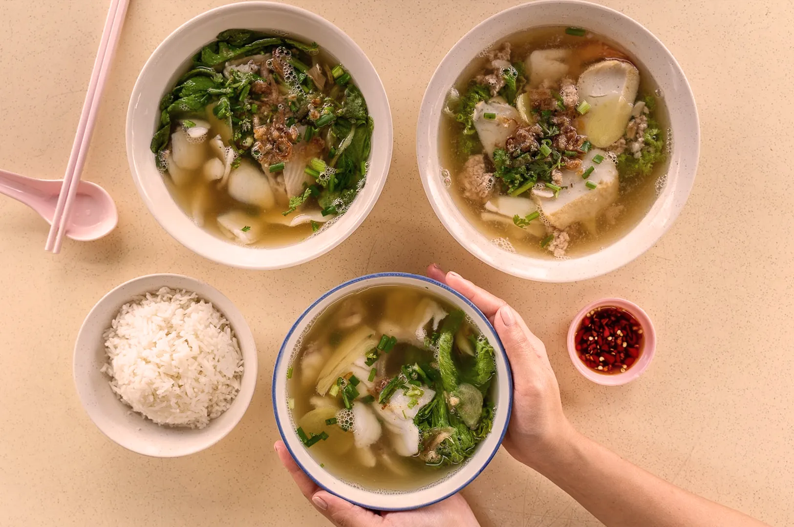 Three bowls of soup with greens and tofu, surrounded by a bowl of rice, pink chopsticks, and a small dish of red chili. Hands are holding one bowl. The scene evokes warmth and comfort.