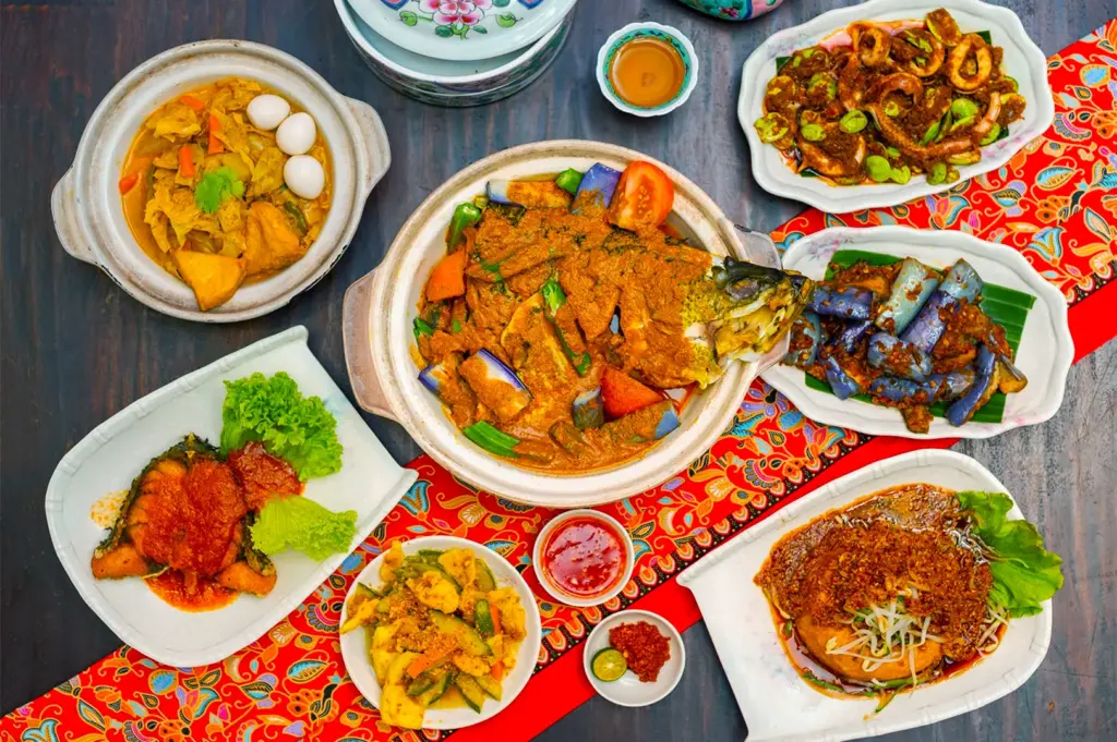 A vibrant spread of Malaysian dishes on a wooden table, featuring curry fish head, spicy chicken, roasted vegetables, and side sauces, exuding a festive and appetizing atmosphere.