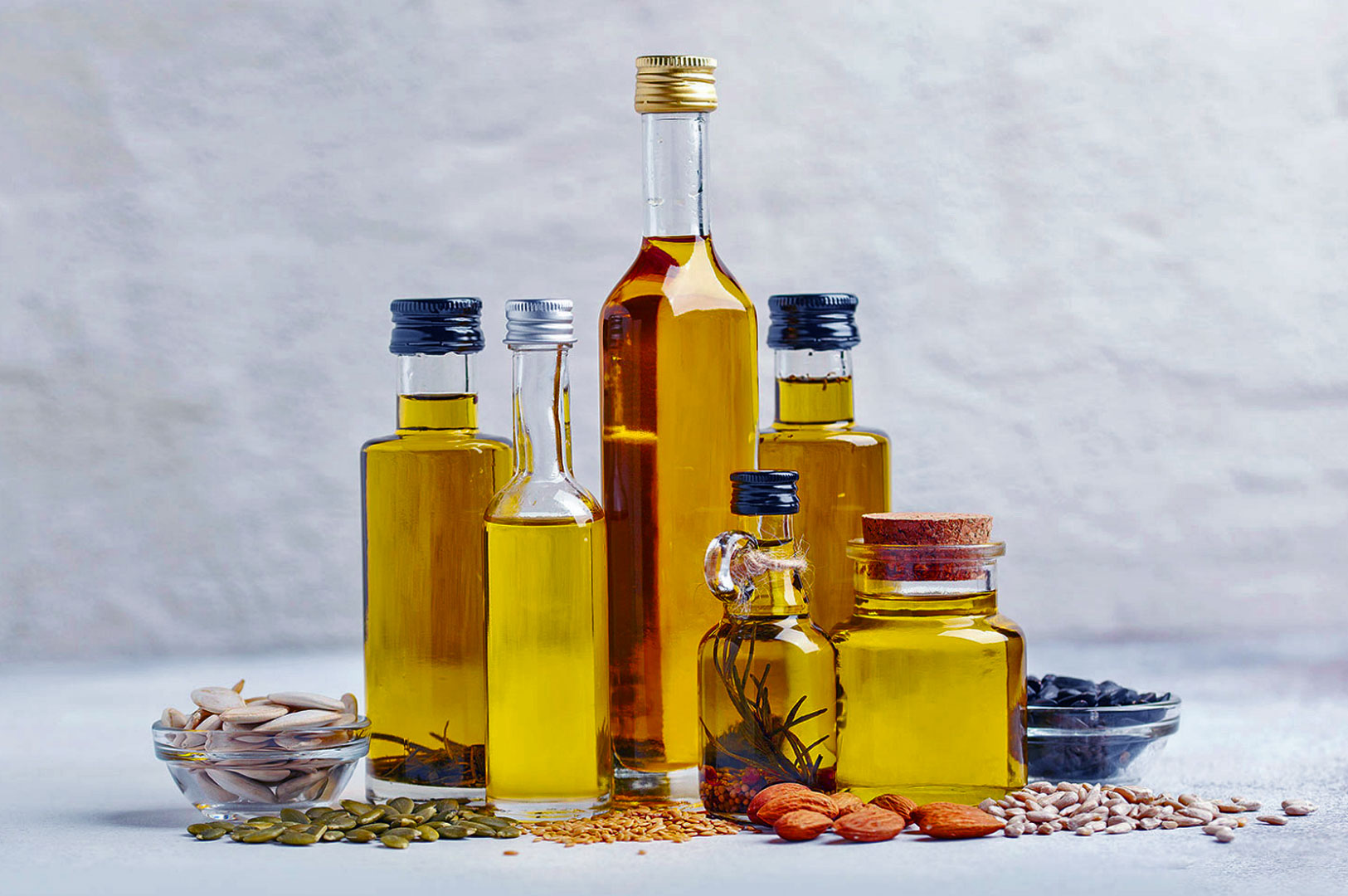 Assorted glass bottles of various oils, surrounded by seeds and nuts, are displayed on a light background. The scene conveys freshness and variety.