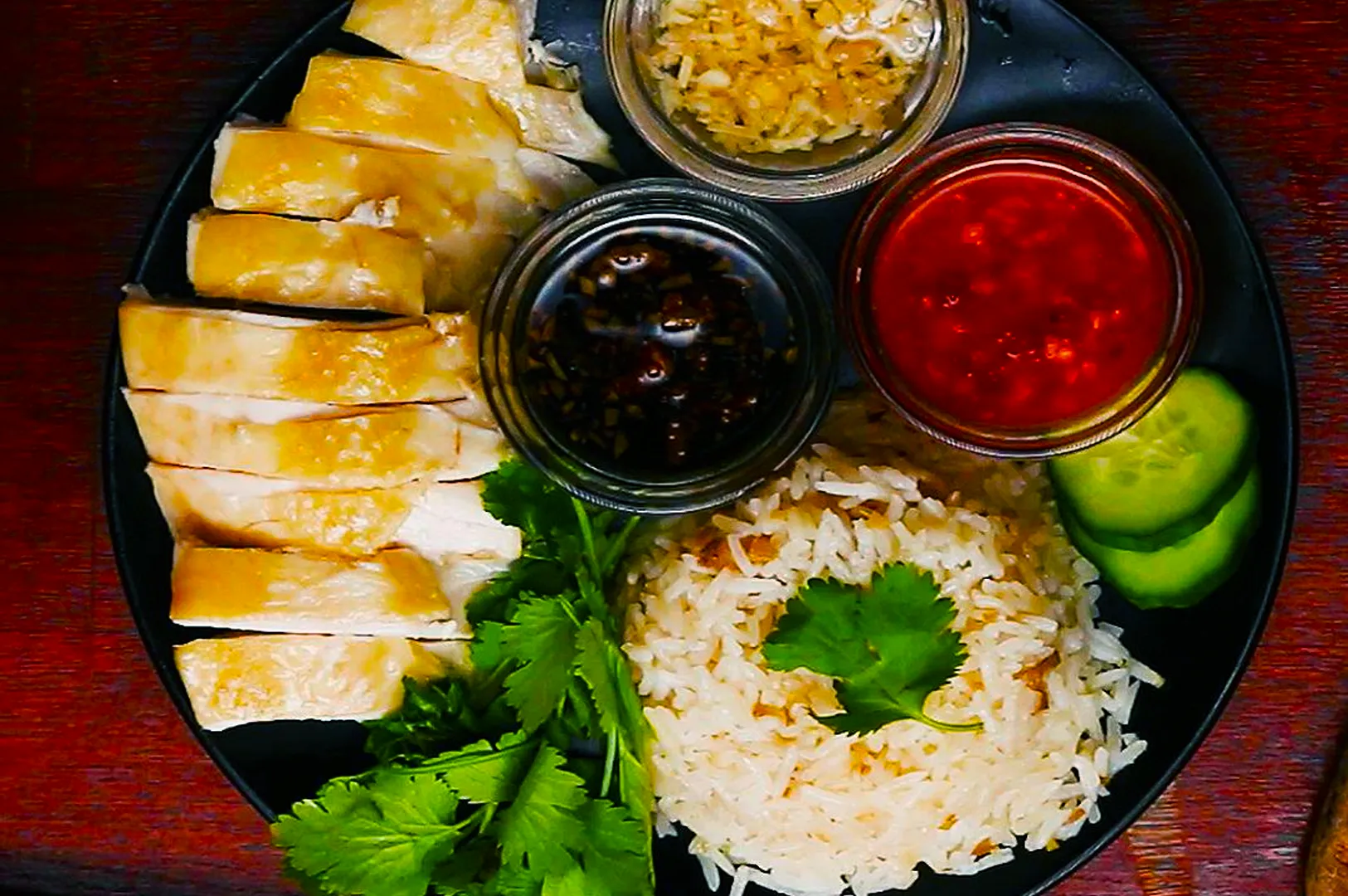 A black plate with sliced chicken, fragrant rice with cilantro, cucumber slices, and three sauces in jars: one red, another dark, and minced garlic.
