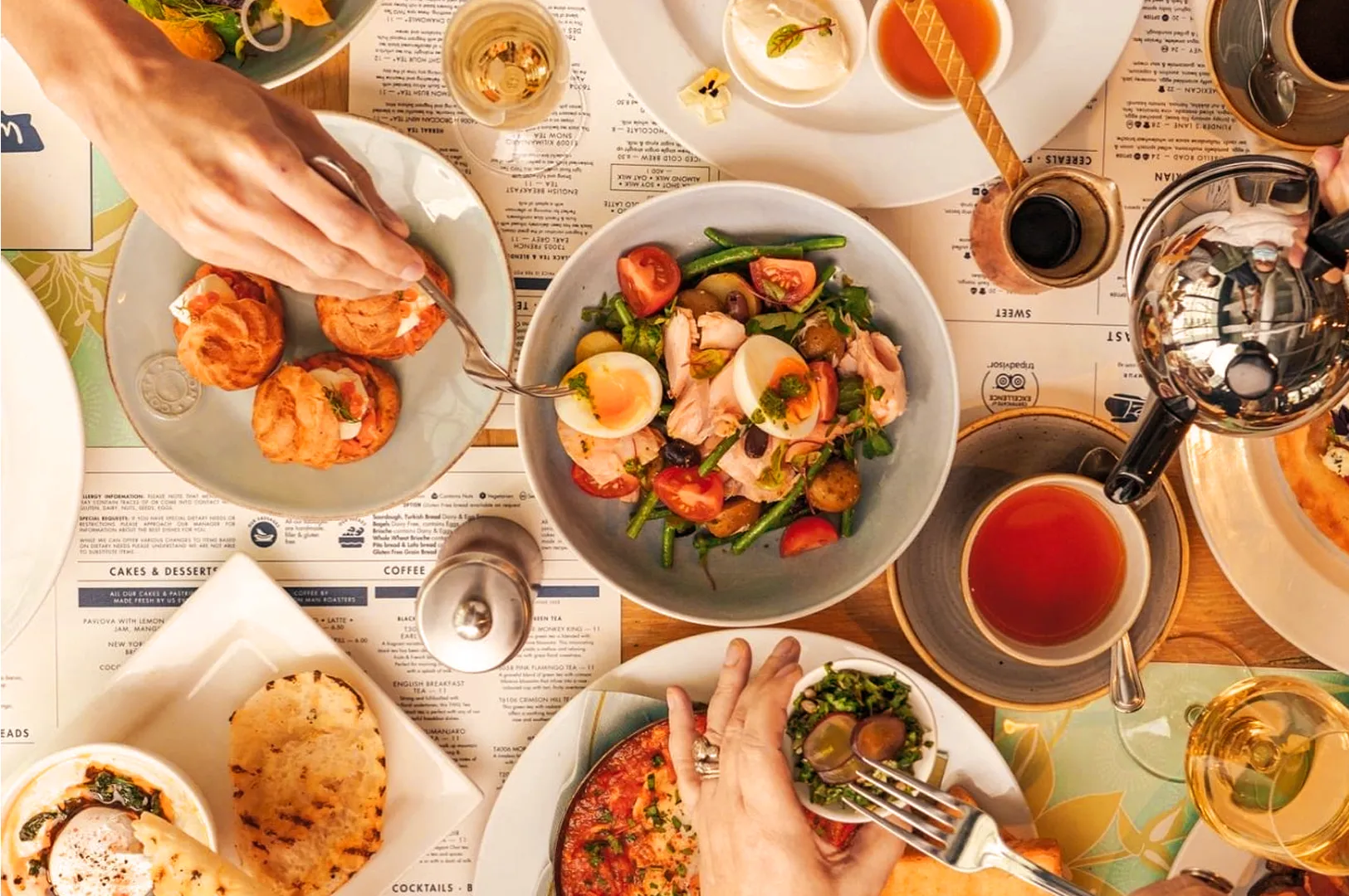 A vibrant table spread with a colorful salad, deviled eggs, pastries, and pizza. Hands reach in serving and pouring drinks, creating a lively dining setting.
