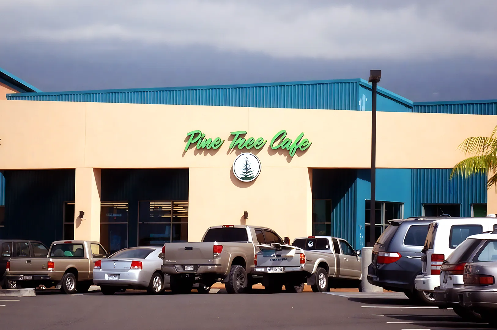 Parking lot in front of a cafe with beige walls and turquoise roof. The sign reads "Pine Tree Cafe." Several cars are parked outside.