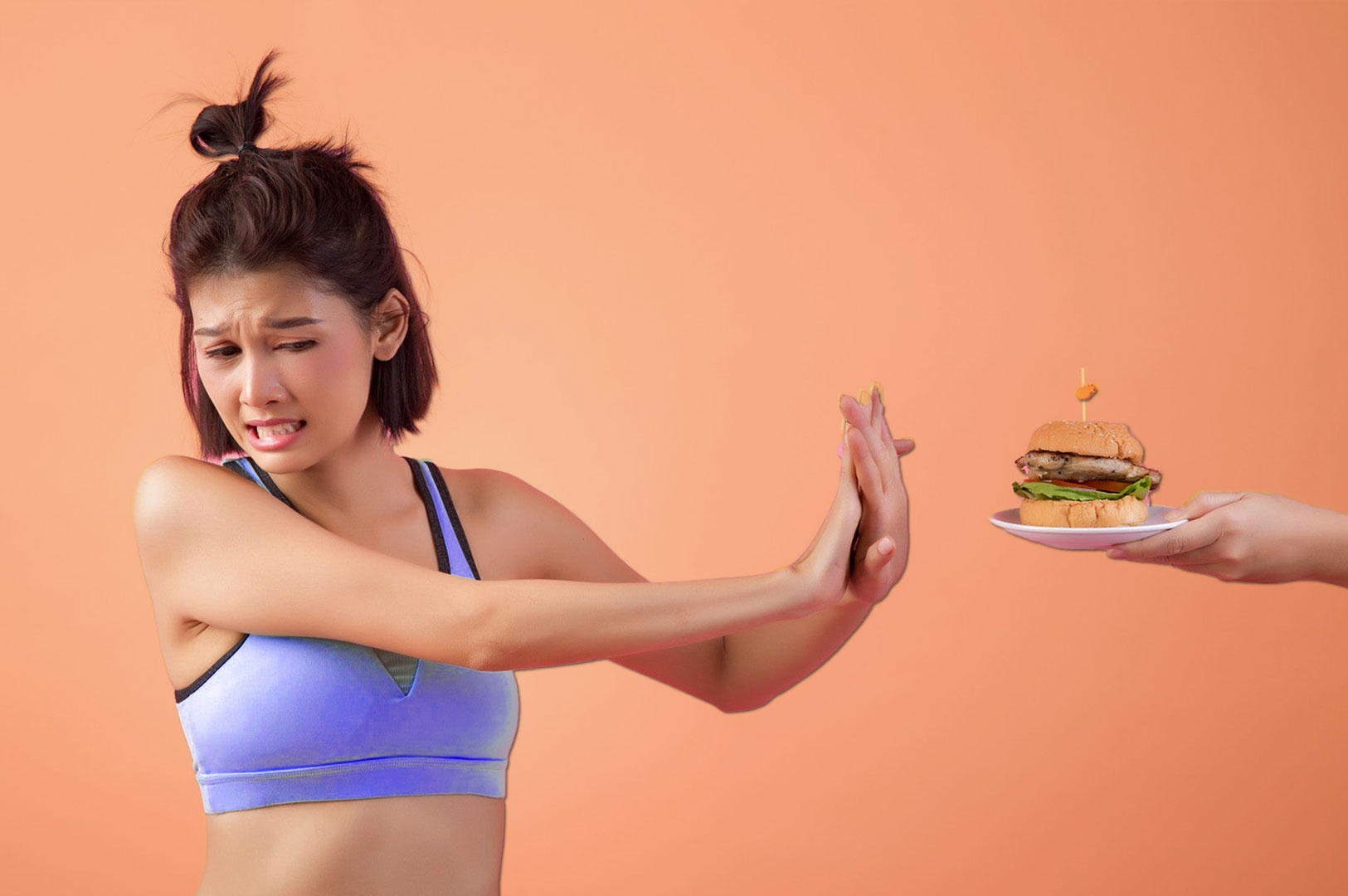 A woman in a sports bra, with a distressed expression, extends her hand to reject a burger on a plate against a peach background; tone conveys conflict.