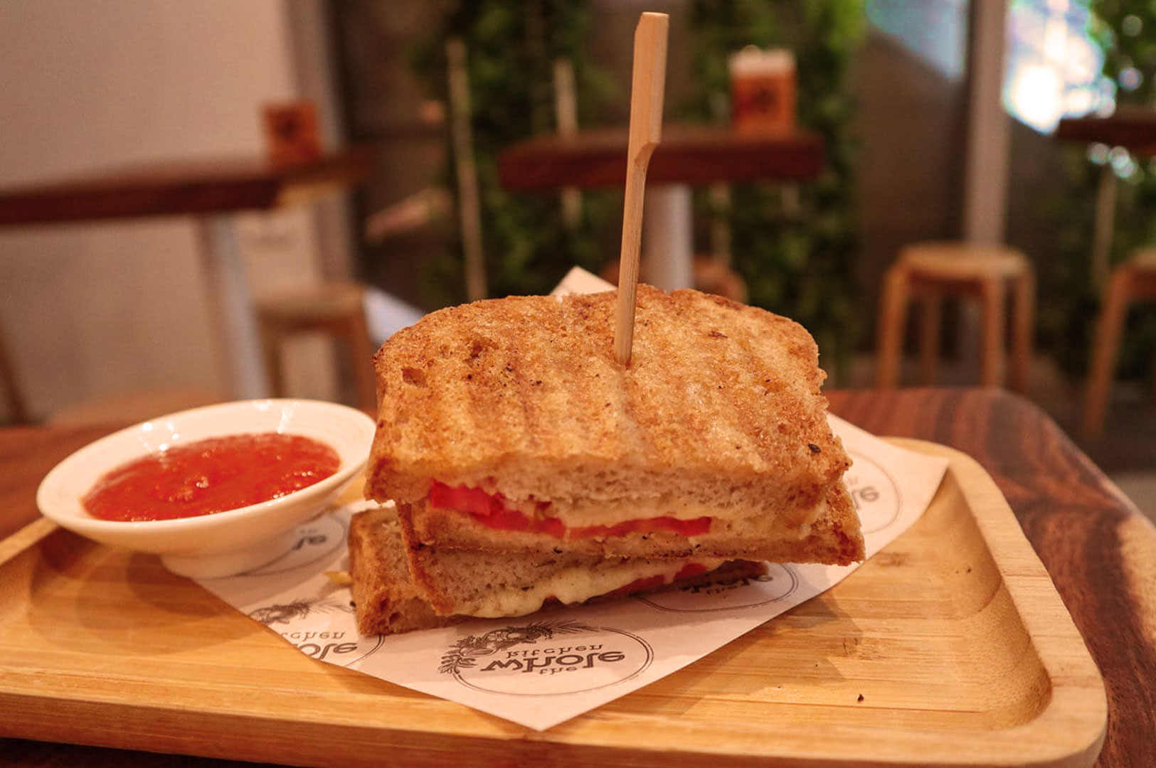 Grilled cheese sandwich with tomatoes stacked on a wooden tray, skewered with a stick. Served with a bowl of red sauce. Rustic cafe background.