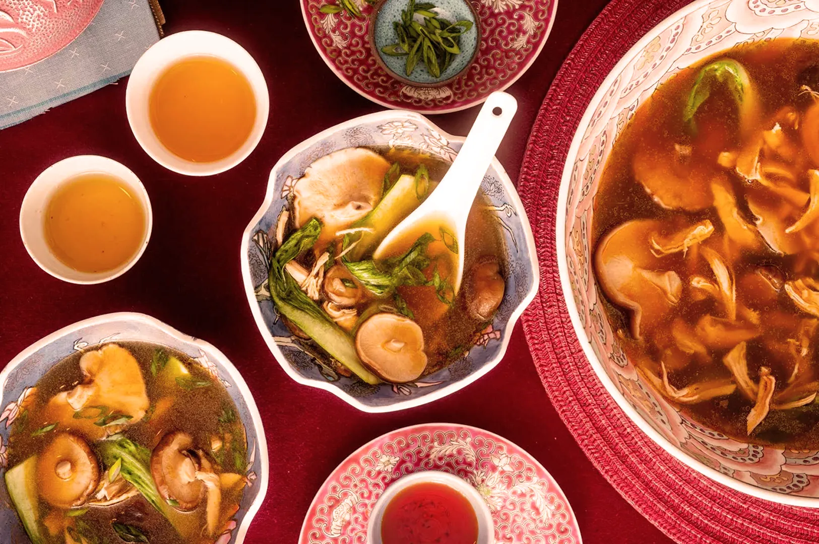 A vibrant spread of Asian cuisine on red tablecloth, featuring bowls of soup with mushrooms and greens, tea cups, and a dish of green garnish, evoking warmth.