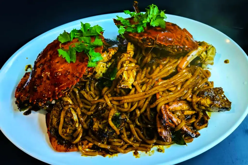 A white plate of spicy crab noodles garnished with fresh cilantro. The vibrant dish features whole crabs, thick noodles, and a rich, dark sauce.