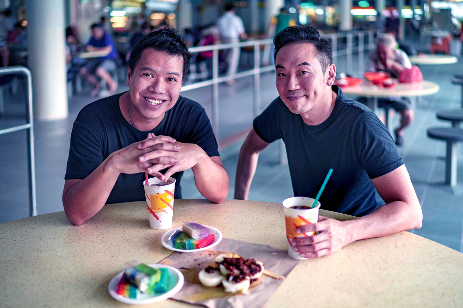 Two people sit at a table smiling, each holding a drink. Plates of colorful desserts and snacks are in front of them in a casual indoor setting.