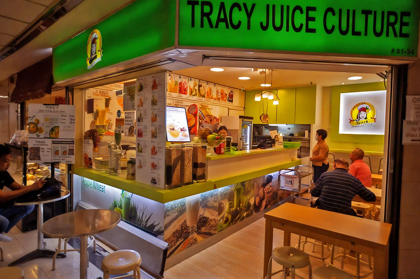 A vibrant juice bar with a bright green sign, "Tracy Juice Culture." Inside, customers sit at tables and the counter is adorned with fresh fruits and menu images.