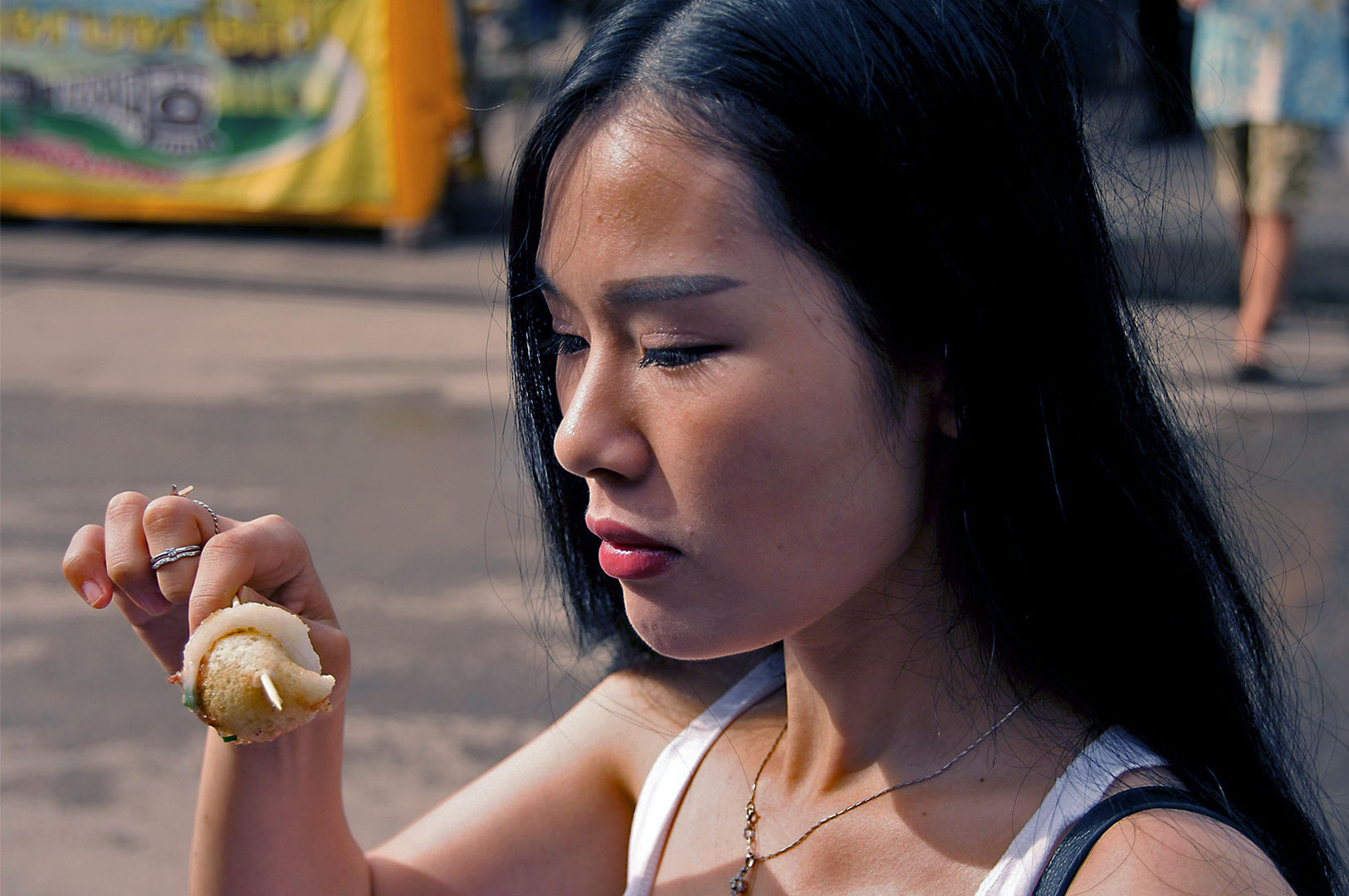 A woman with long dark hair examines a piece of street food on a skewer, looking thoughtful and curious. Blurred market scene in the background.