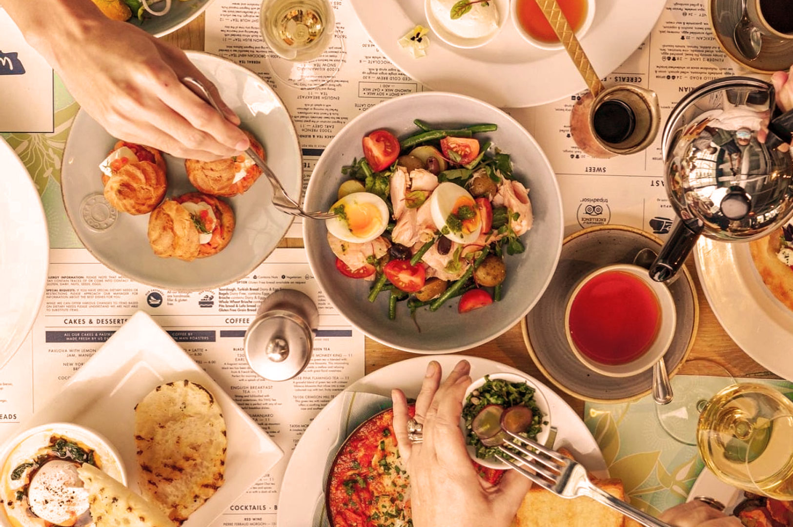 Aerial view of a brunch table with various dishes, including a salad with eggs and tomatoes, pastries, pizza, and tea. Hands reach for food, creating a lively, communal atmosphere.