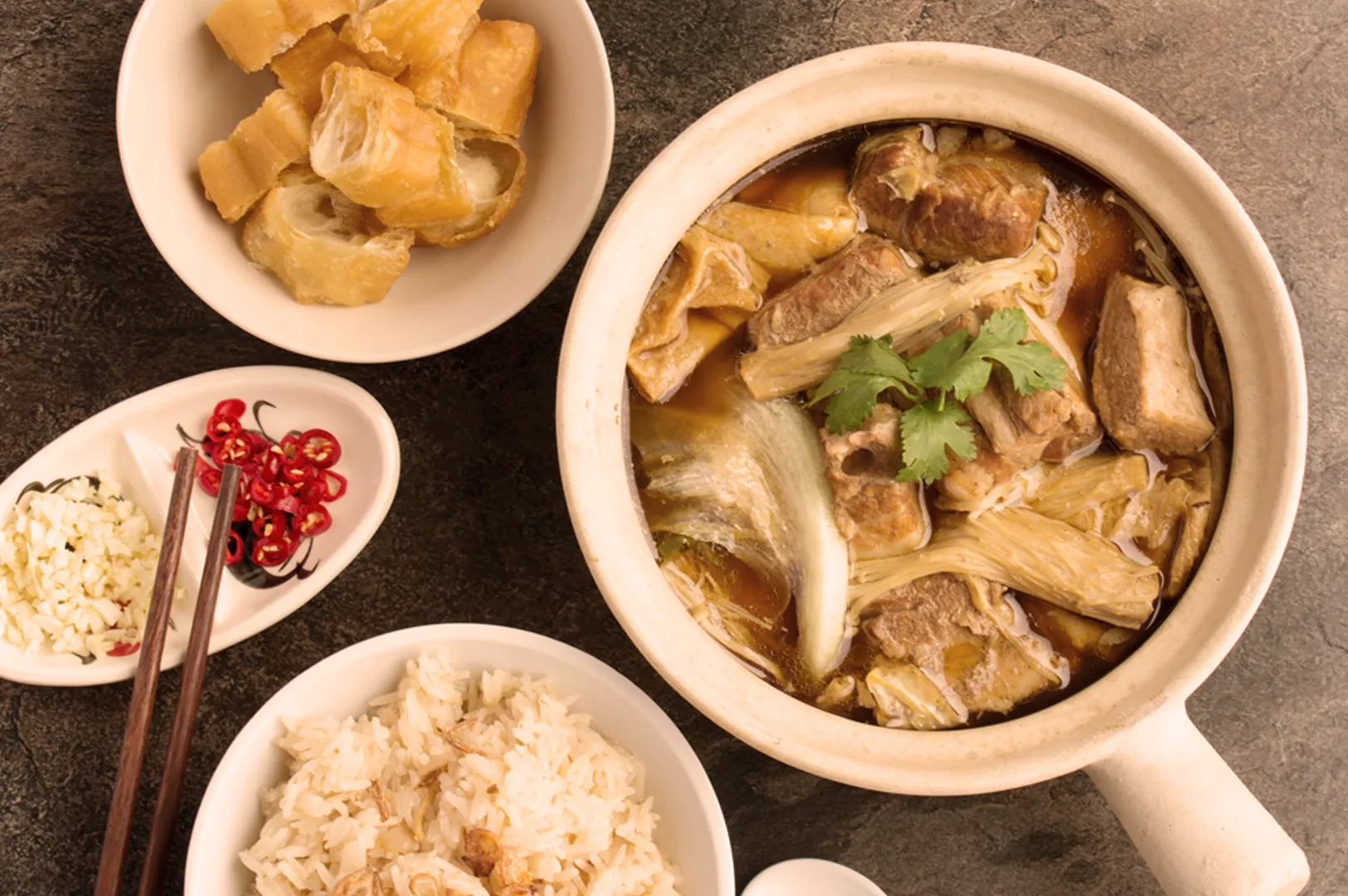 A flavorful Asian clay pot soup with meat, tofu, and cilantro. Served with a bowl of rice, fried dough, and garnished with sliced garlic and chili. Cozy and inviting.