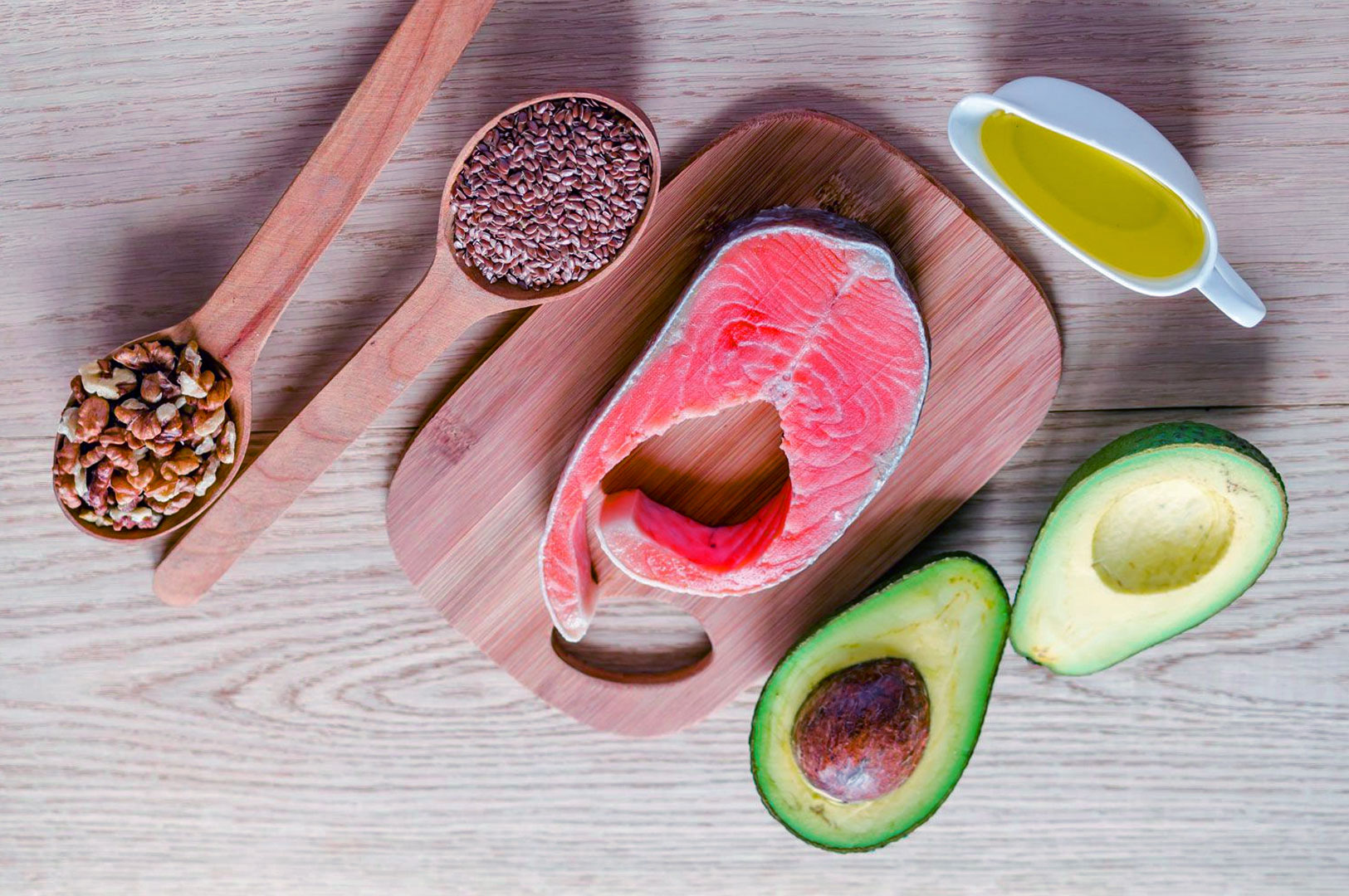 Salmon steak on a wooden board surrounded by flaxseeds, walnuts, avocado halves, and a small jug of olive oil, highlighting healthy fats.