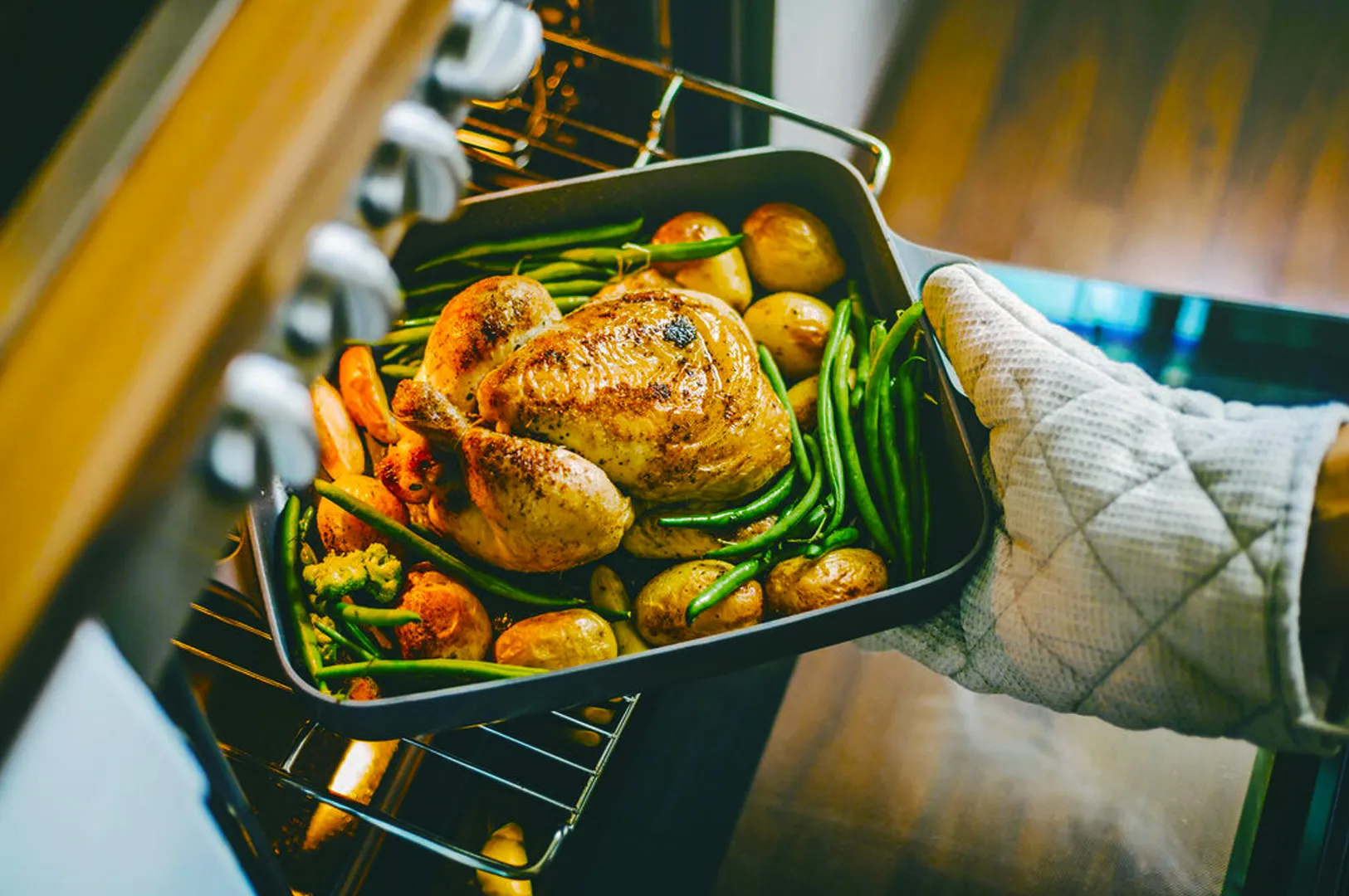 A gloved hand removes a roasted chicken tray with potatoes, green beans, and carrots from an oven. The scene suggests warmth and a home-cooked meal.