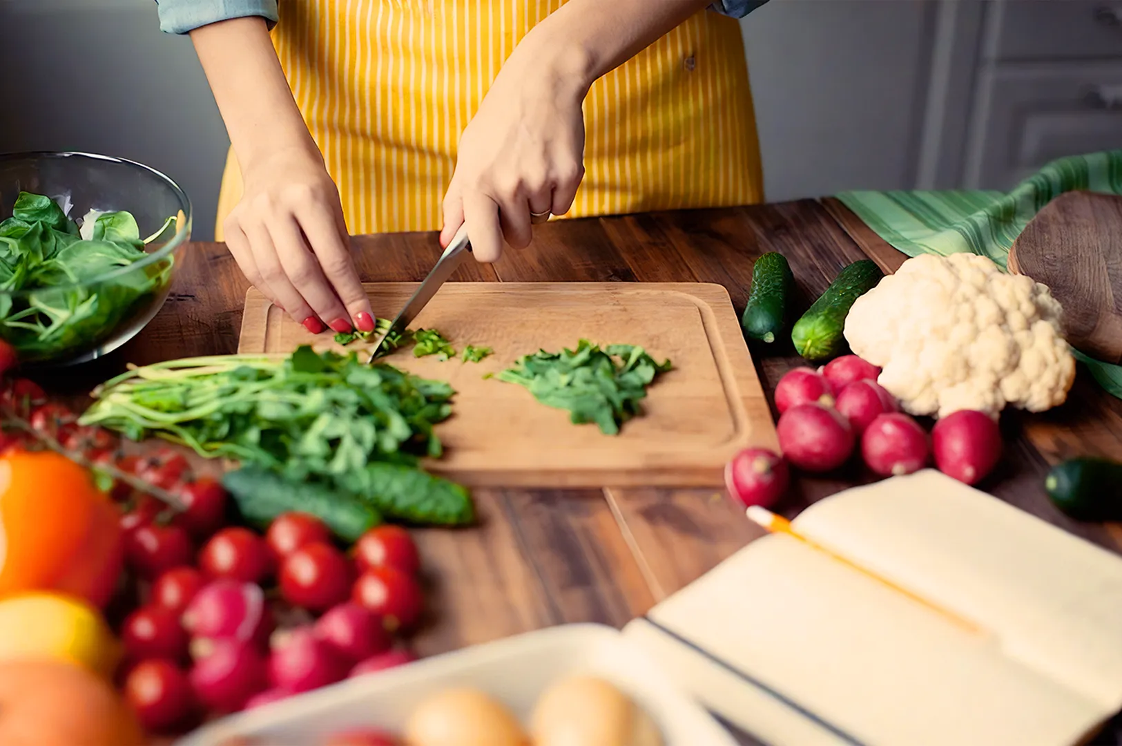 Hands chop leafy greens on a wooden board in a bright kitchen. Vibrant vegetables, including radishes and cauliflower, are scattered nearby.