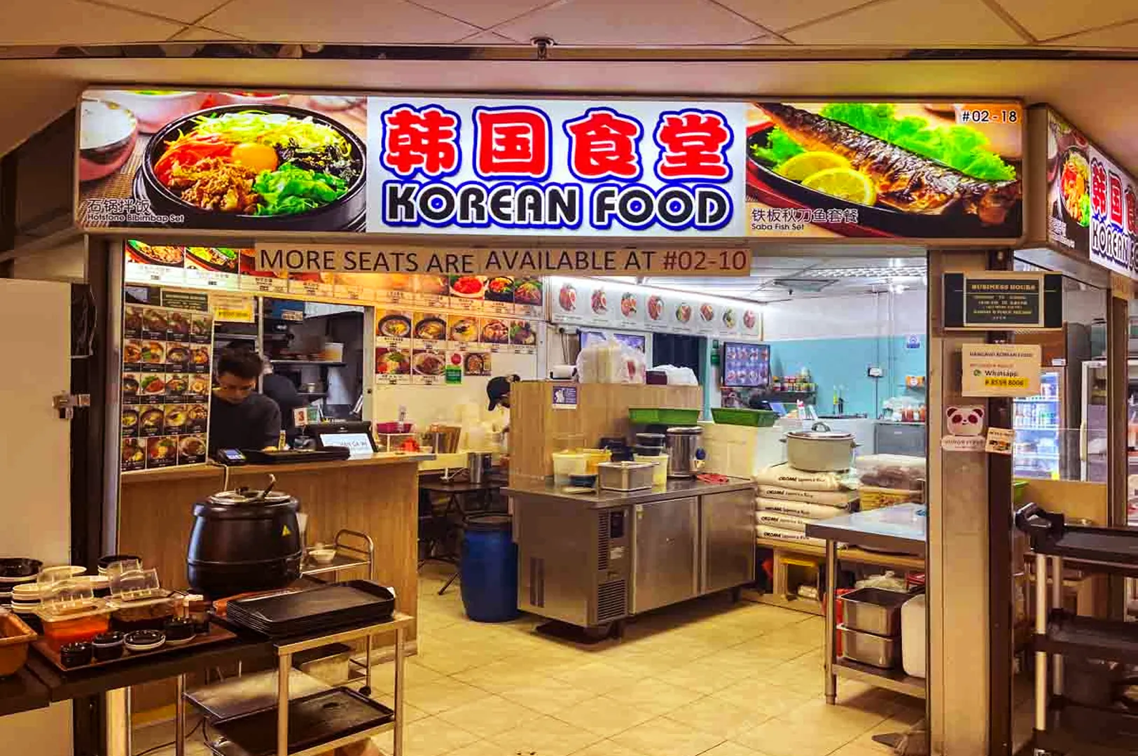 Brightly lit Korean food stall with colorful signage and food images above the counter. A staff member works inside. Cozy and inviting atmosphere.