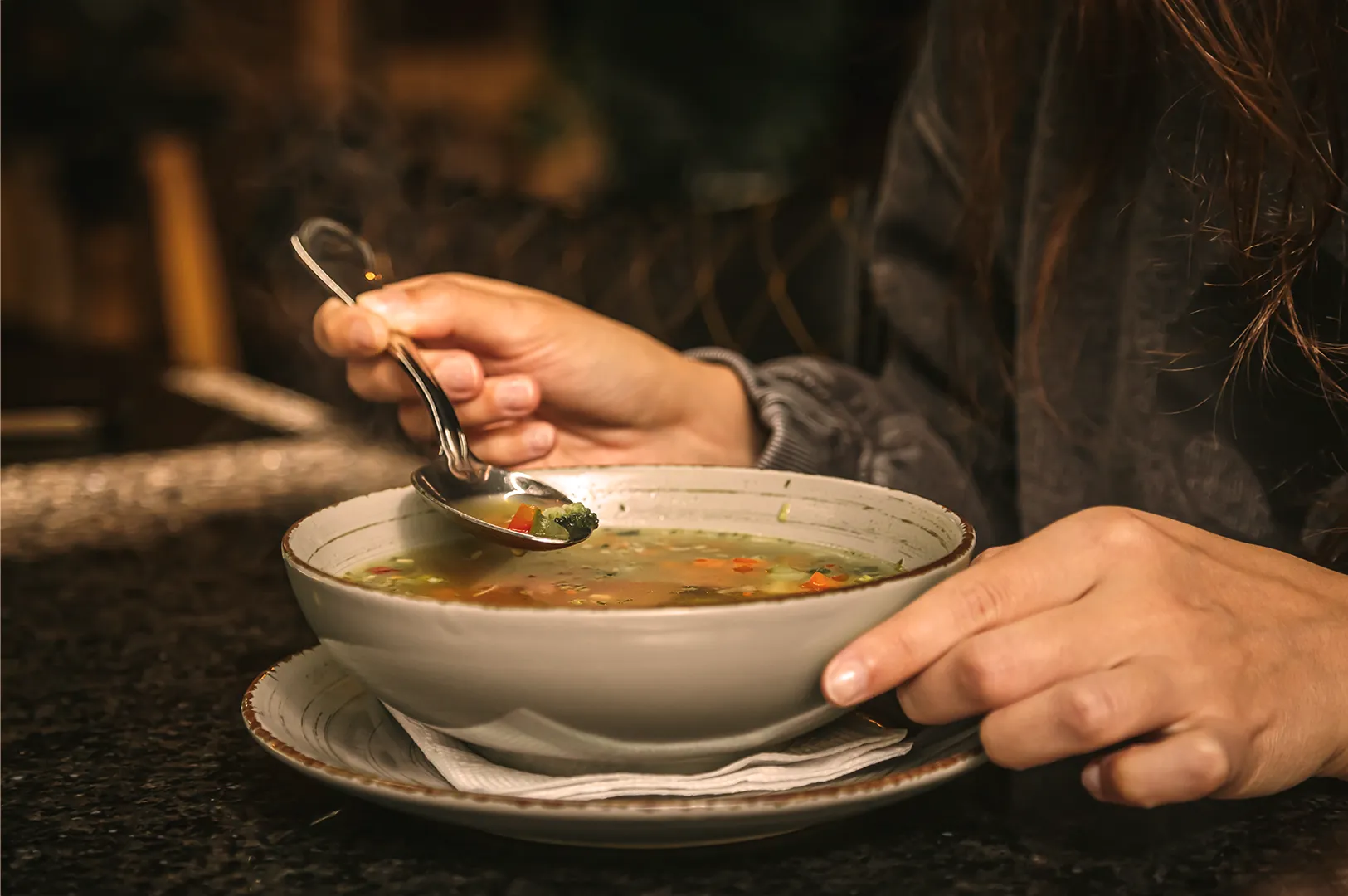 A person holds a spoon above a bowl of vegetable soup, ready to eat. The warm lighting and cozy setting suggest a comforting meal.