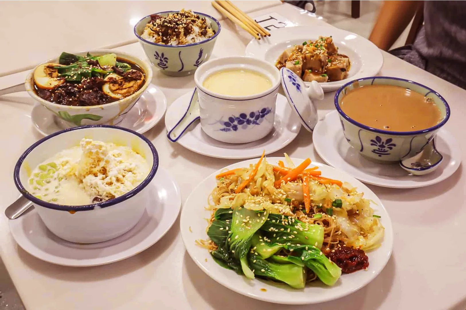 A colorful spread of Chinese dishes on a table, including stir-fried noodles, chicken rice, steamed tofu, egg custard, tapioca dessert, and a cup of tea.