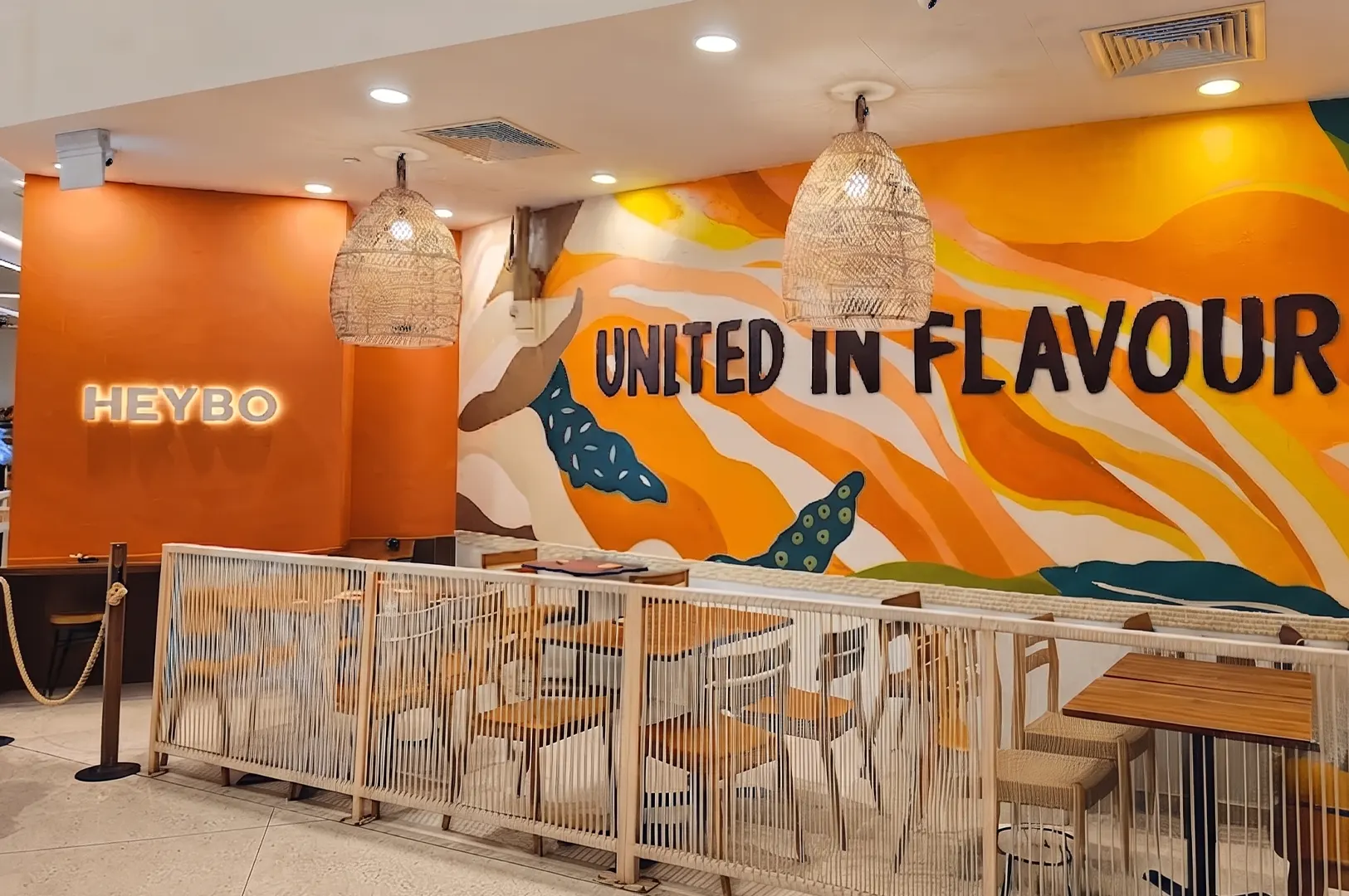 Modern restaurant interior featuring wooden dining tables, woven light fixtures, and a vibrant orange wall mural that reads "UNITED IN FLAVOUR".