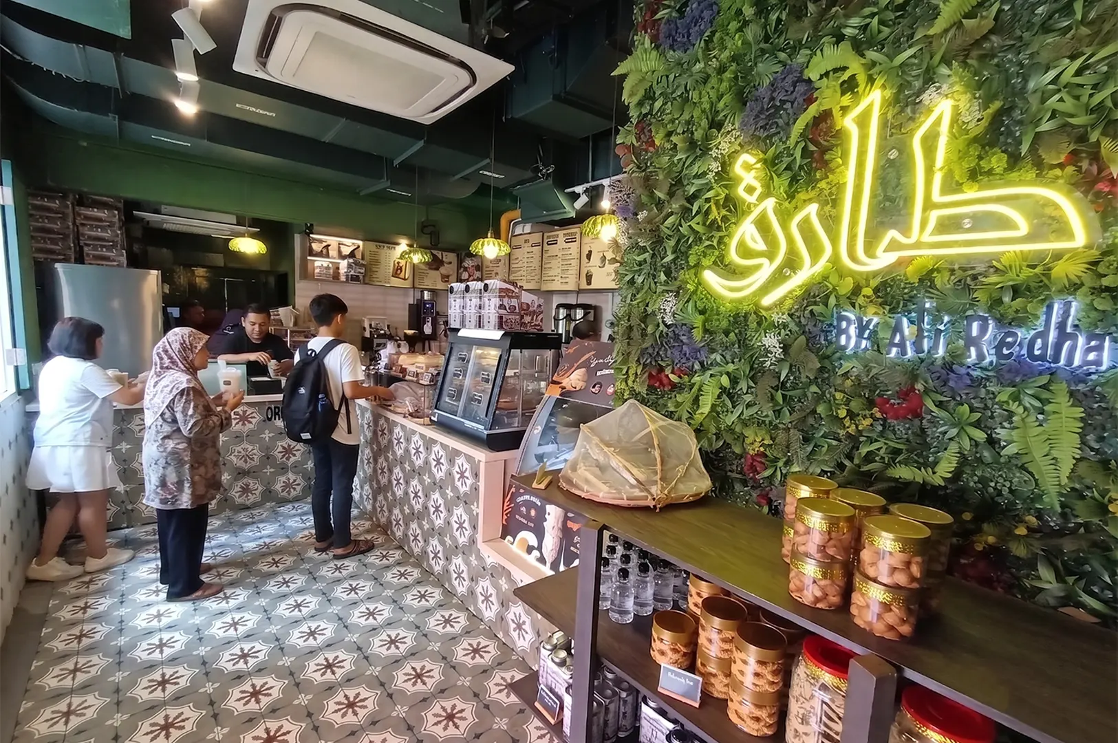 Interior view of the cafe Tarik by Ali Redha, featuring a green wall with neon signage, decorative tiled flooring, and customers ordering at the counter.