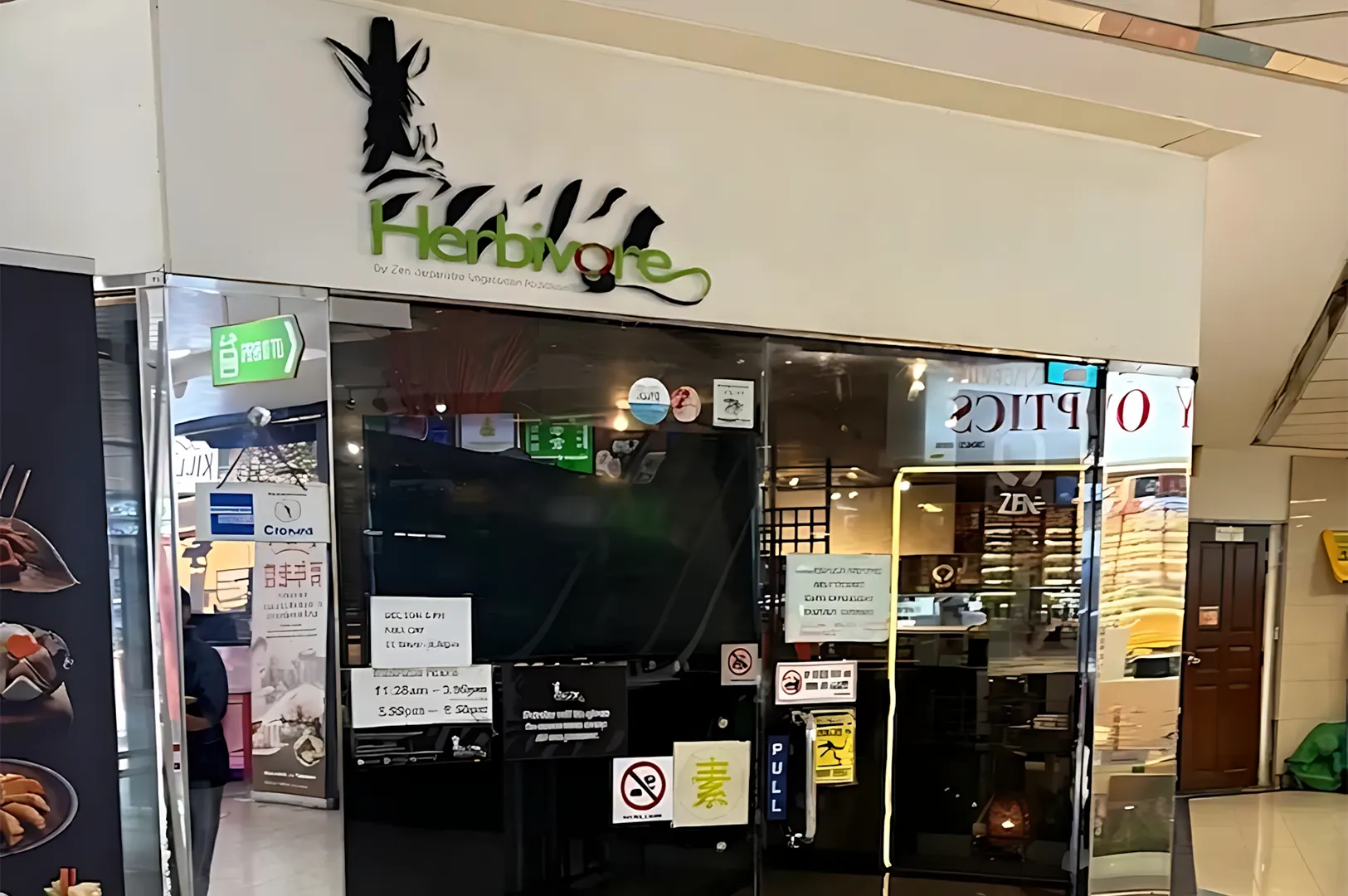 The dark glass storefront entrance of Herbivore restaurant with a white logo featuring a stylized zebra profile above the green text.