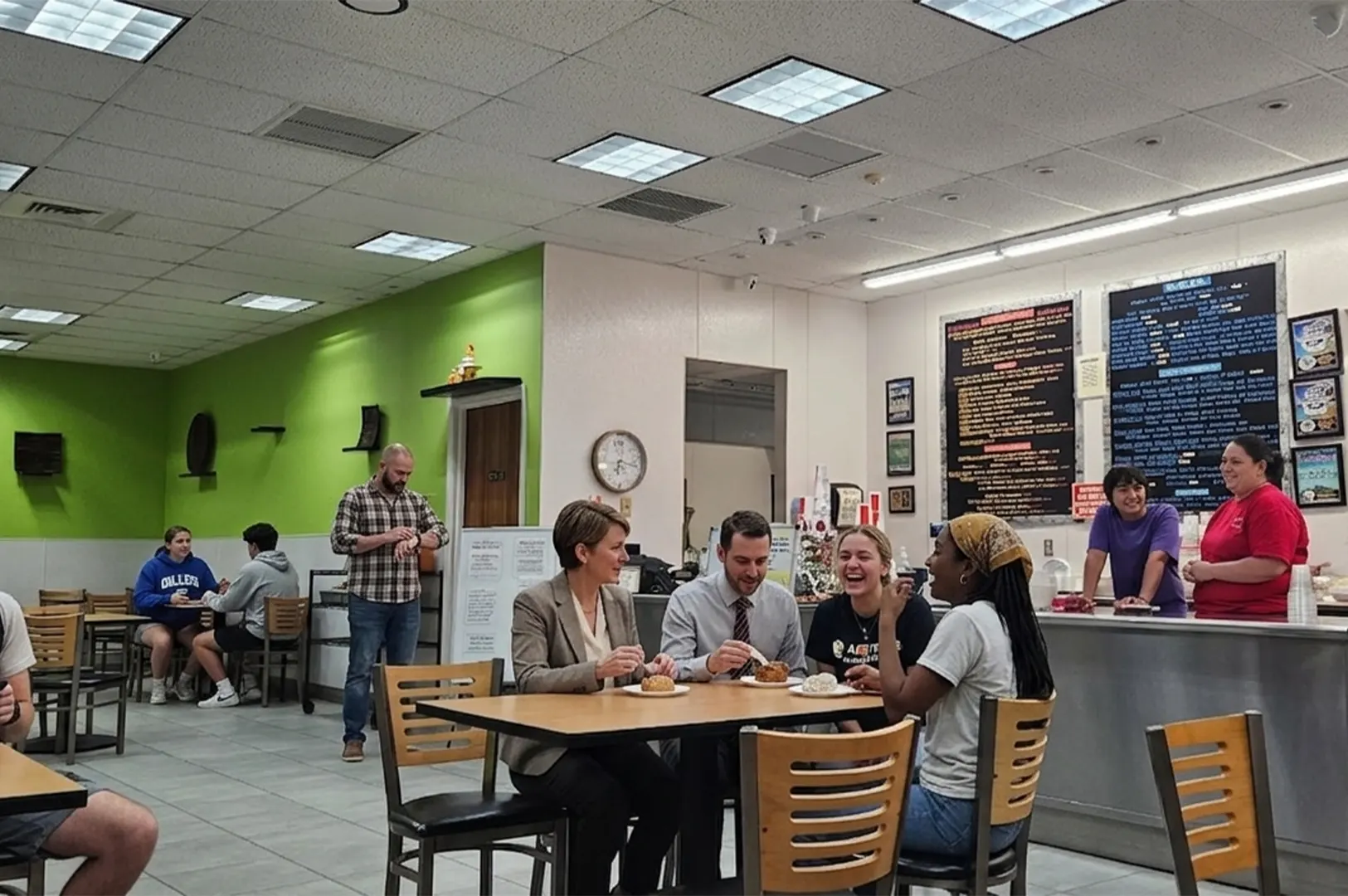 Customers dining and laughing at a table inside Pine Tree Cafe with a service counter and menu boards in the background.