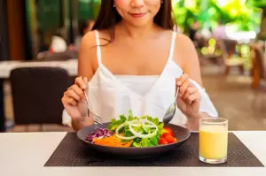 A woman in a white top is smiling slightly while sitting at a table with a fresh salad and a glass of juice. The background is a blurred outdoor setting.