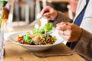 Person eating a healthy salmon grain bowl with leafy greens and vegetables at a sunlit wooden table.