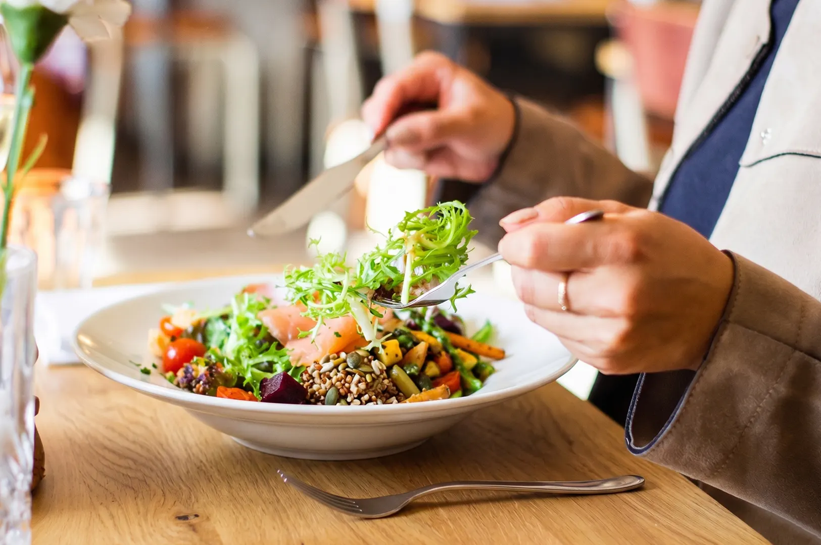 Person eating a healthy salmon grain bowl with leafy greens and vegetables at a sunlit wooden table.
