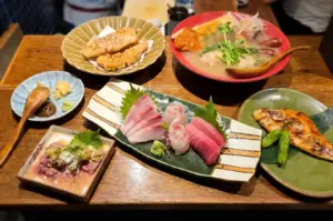 A variety of food on a wooden table, including plates of sashimi, fried tempura, grilled fish, and a bowl of soup against a blurry background.
