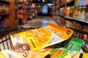 Shopping cart in a grocery aisle with colorful snack packages, including a prominent orange mango bag. Shelves are filled with various items. Warm lighting.